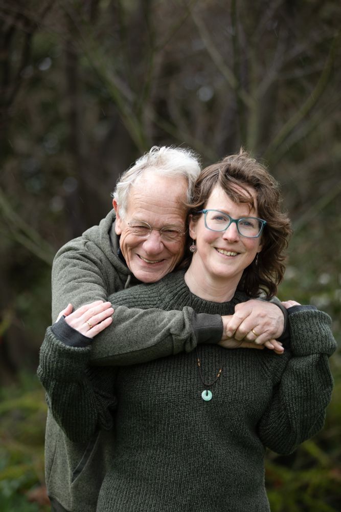 Photo shows me and my husband - a white couple - looking into the camera and smiling. My husband stands behind me hugging me and I have my hand on his arm. We are both wearing green and there is a natural green background. 