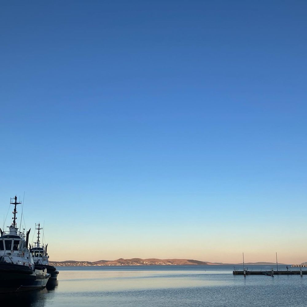 Photo taken at the Nipaluna/Hobart waterfront shows two tug boats to the left and a pier to the right. The water of the River Derwent is calm, sun shines on the Eastern shore, and there is a big blue sky above.