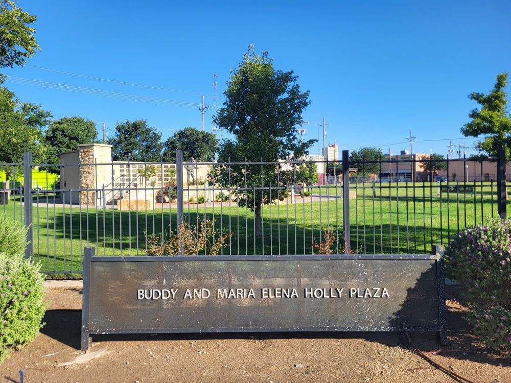 A metal sign in front of a metal fence says, "Buddy and Maria Elena Holly Plaza." Behind the fence is a stone monument with a bronze statue of musician and Lubbock, Texas-native Buddy Holly.