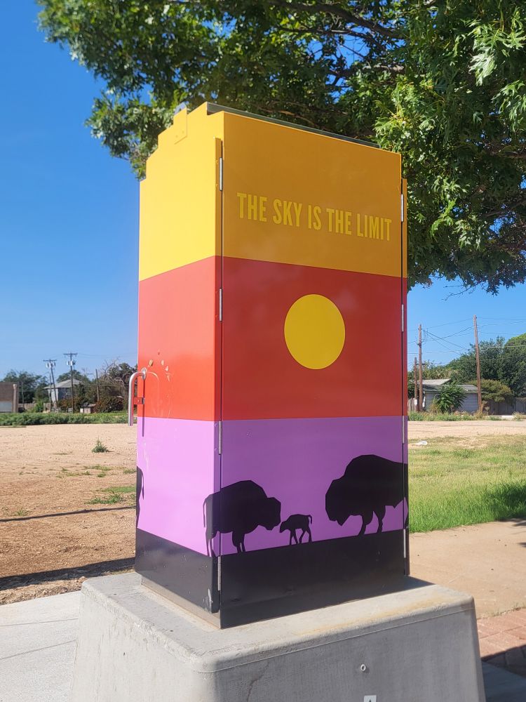 A power box on a pedestal with a mural painted on it. The mural has broad stripes of yellow, red, light purple, and black. Text says "the sky is the limit" in the yellow stripe with a yellow sun on the red stripe and black bison on the light purple stripe. Immediately behind the power box are some tree leaves. A red plot of land sits behind it with some rundown houses in the distance.
