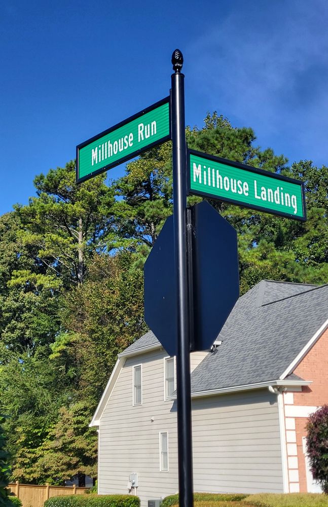An intersection with a stop sign and two roads signs above it. The road signs say: Millhouse Run and Millhouse Landing. There is a house in the background with some large trees, too.