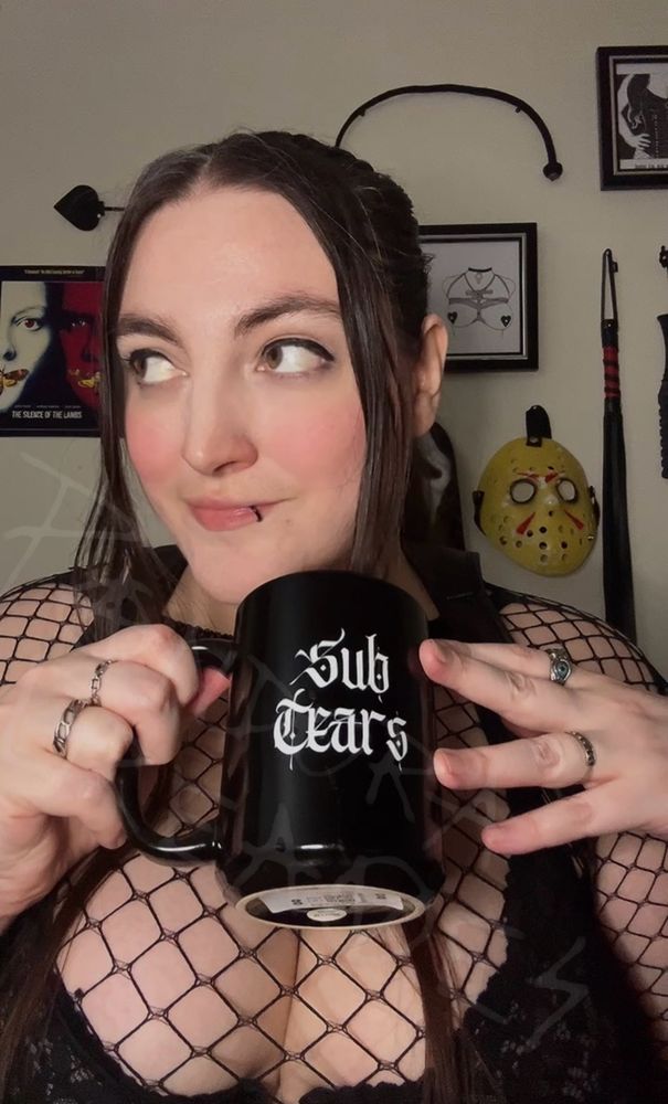 Miss Pandora Greaves, a queer, thick, curvy, big breasted genderfluid God / Goddess with brown hair, hazel eyes, and a lip ring is seen close up, looking off to Their right with a playful smirk while holding a cup that reads “sub tears”. They are wearing a black fishnet top and black lace bra and the cleavage from Their huge tits is very visible.

The wall is adorned with BDSM art by RIP Bambi, posters for the horror movies Silence of the Lambs, John Carpenters Halloween, and Evil Dead, a mask from Friday the 13th And Scream, as well as implements of pain such as a crop, single tail whip, paddle, and floggers for impact play. 
