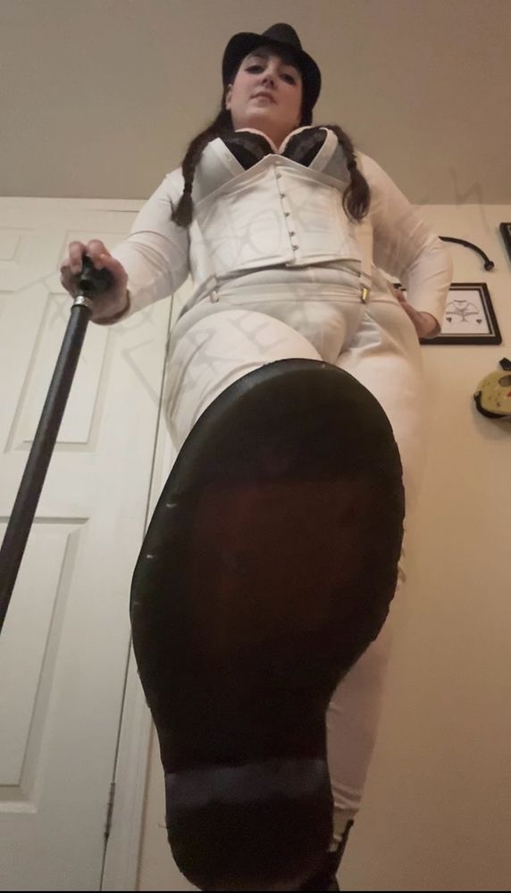 Miss Pandora Greaves, a queer, thick, curvy, big breasted genderfluid God / Goddess with brown hair, hazel eyes, and a lip ring is dressed in black Doc Martens, a black fedora, black bra, white overalls (which the black bra is busting out of), a white corset, and a cod piece held up by suspenders. They are leaning on a dark wooden cane with a skull as the handle and Their right foot is forward so the bottom to Their boot is in your face. They’re dressed as Their own version of a Droog from A Clockwork Orange. 

The wall is adorned with BDSM art by RIP Bambi, posters for the horror movies Silence of the Lambs, John Carpenters Halloween, and Evil Dead, a mask from Friday the 13th And Scream, as well as implements of pain such as a crop, single tail whip, paddle, and floggers for impact play. 
