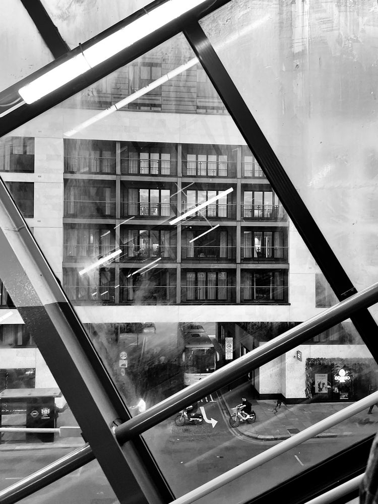 A black and white photo through the window of a London street with high rise building, bus, and bike rider.