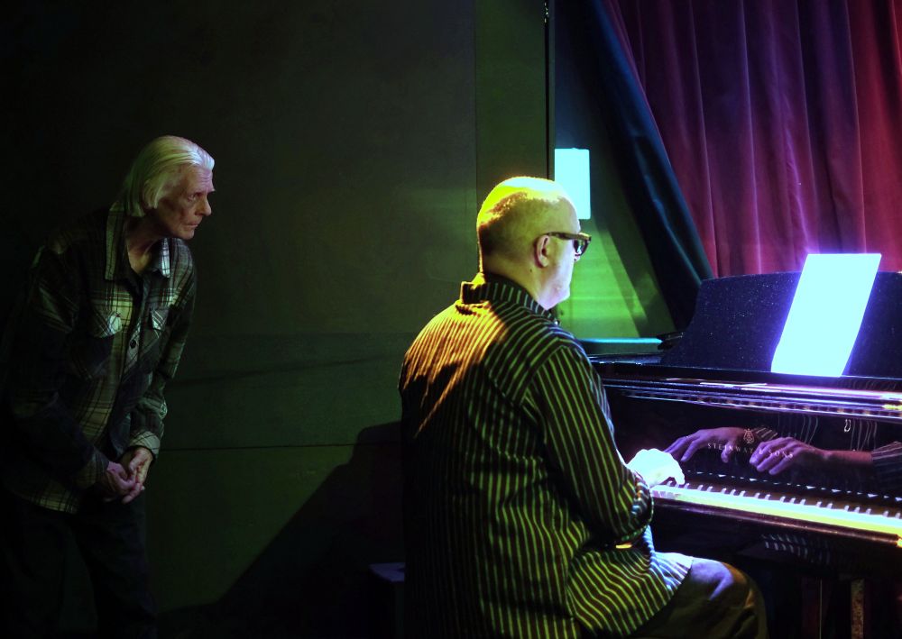 En man sitter vid en flygel och spelar. En äldre herre står bakom honom och spänner blicken på notbladet.