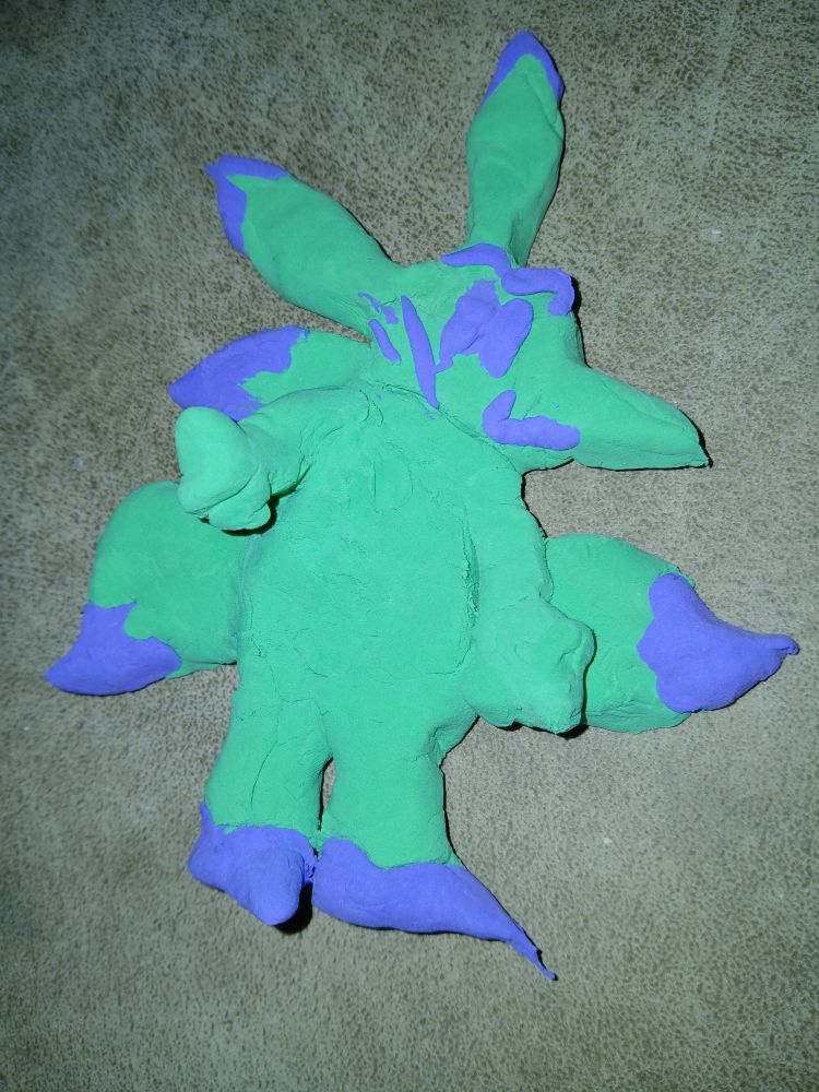 Image of a clay representation of a kitsuna.
It resembles an anthropomorphic fox.
It has two ovular ears without openings, and three tails.
There is an accented purple on the feet, tails, and ears, along with some streakmarks of the same accent on their cheek.