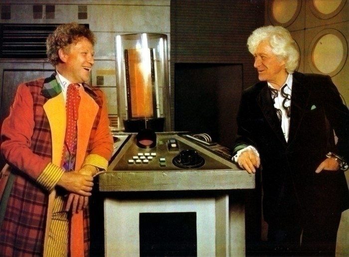 Colin Baker and Jon Pertwee grinning at each other, leaning against the TARDIS console.