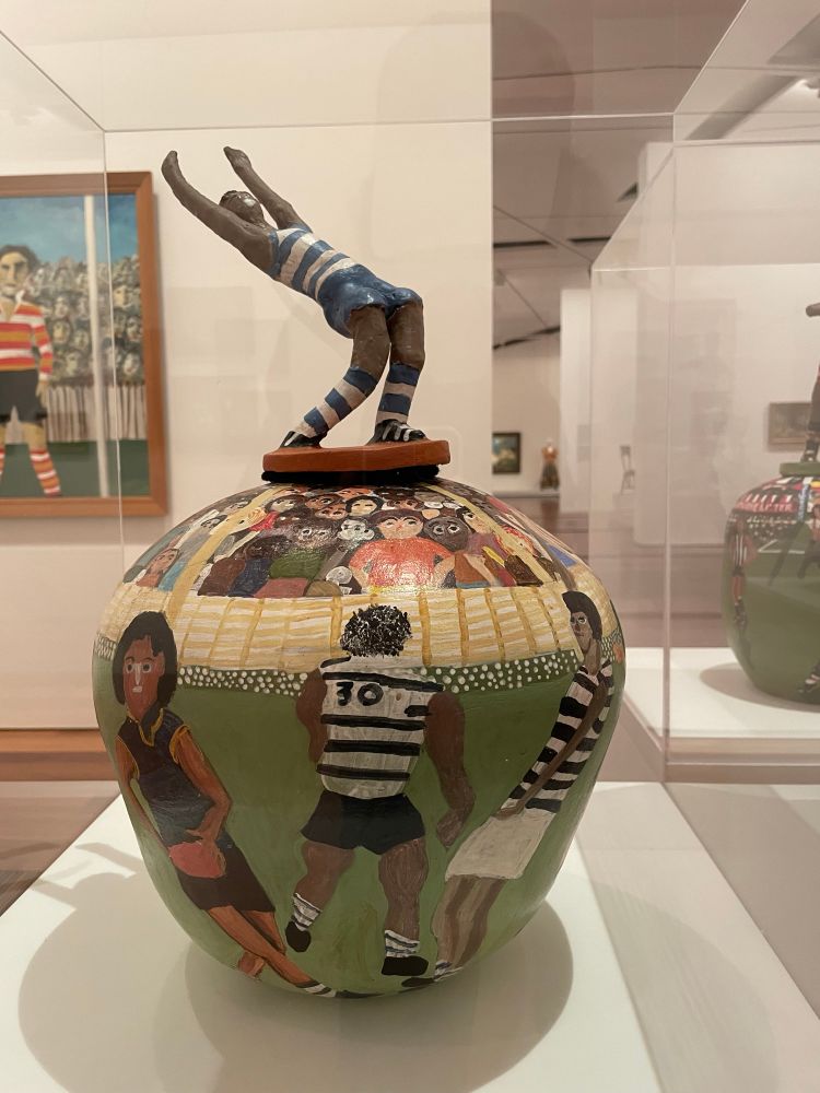 Photograph of a terracotta pottery work in a gallery display case. The vessel is painted with images of a football match with Graham Polly Farmer, who also sits atop. It is by artist Rahel Kngwarria Ungwanaka.