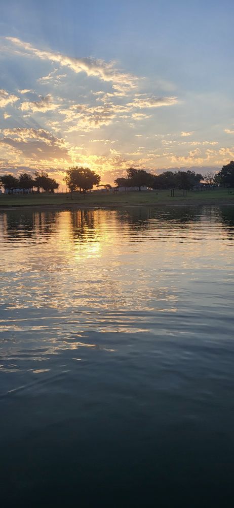 Sunrise with trees in the back and lake water in the front, clouds up top