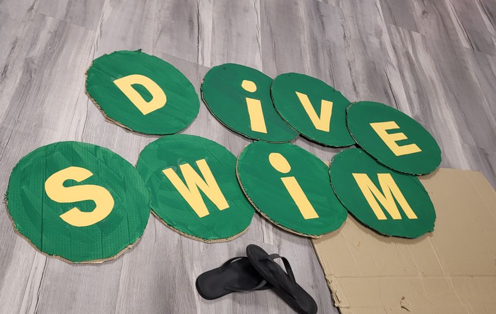 The words dive and swim are in yellow in the center of cardboard circles painted green. Flip flops at the bottom ans a piece of extra cardboard on the bottom right.