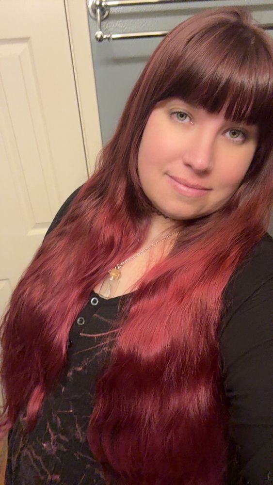 A pale woman with long burgundy hair and green eyes looks up at the camera. She is wearing a black and pink lightning print dress and has a small tired smile. 