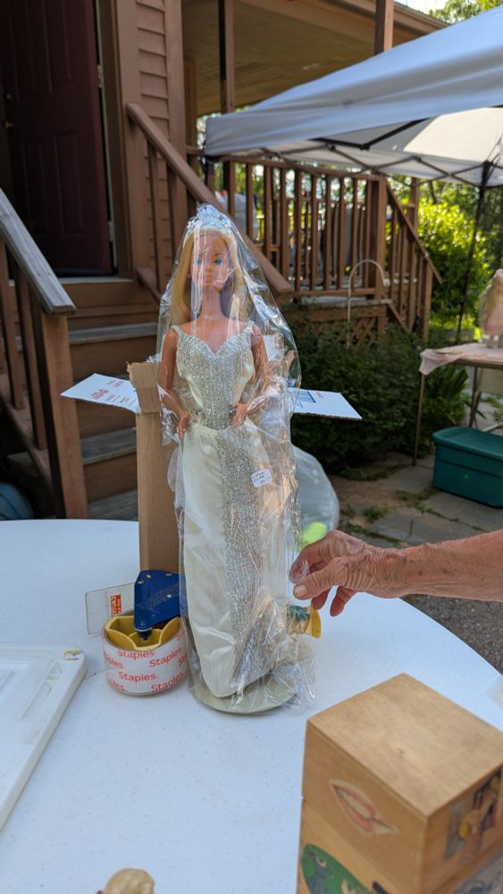 1978 18" Barbie Doll in a white wedding gown wrapped in plastic sitting on a yard sale table. 