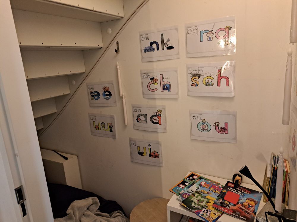 Foto van de onderkant van de trap in de kast, met kussen en letter tekeningen aan de muur, ook het boekenkastje