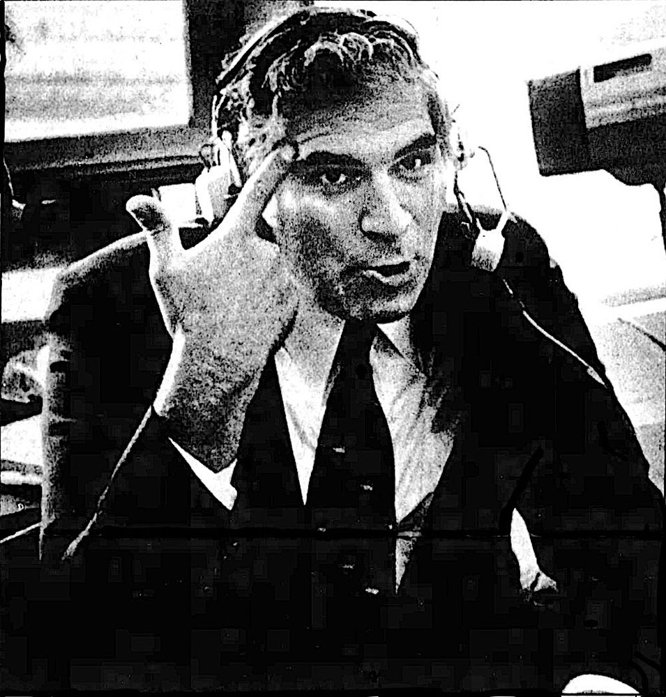 Ron Luciano in jacket and tie, wearing microphone and headset in broadcast booth, pointing to his head.