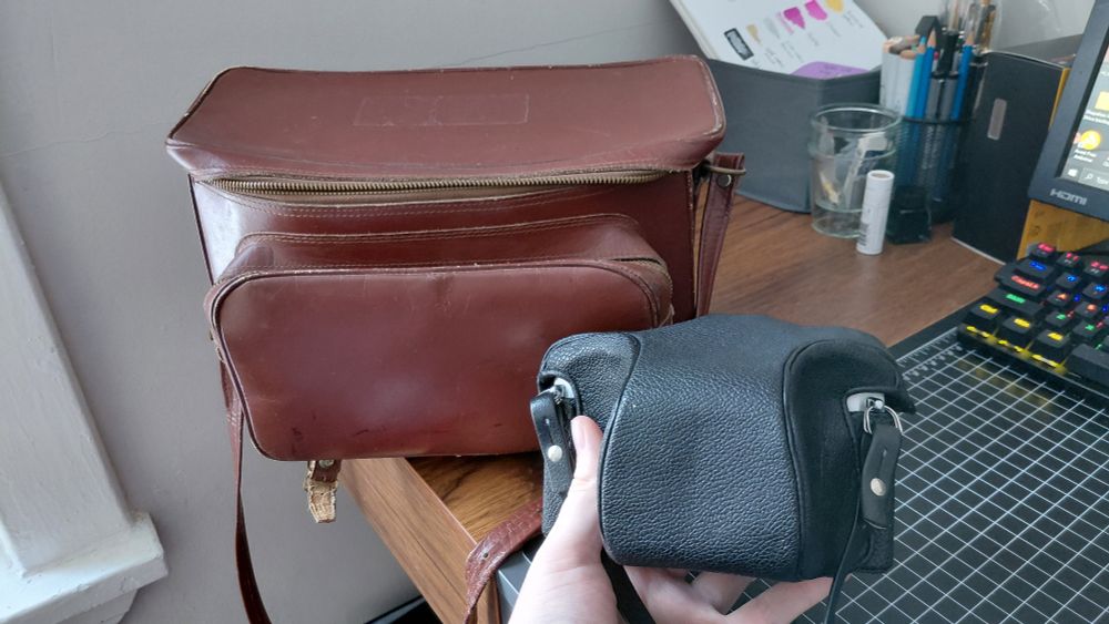 The same camera as before, now in its protective leather cover, next to the leather shoulder strap bag it came with