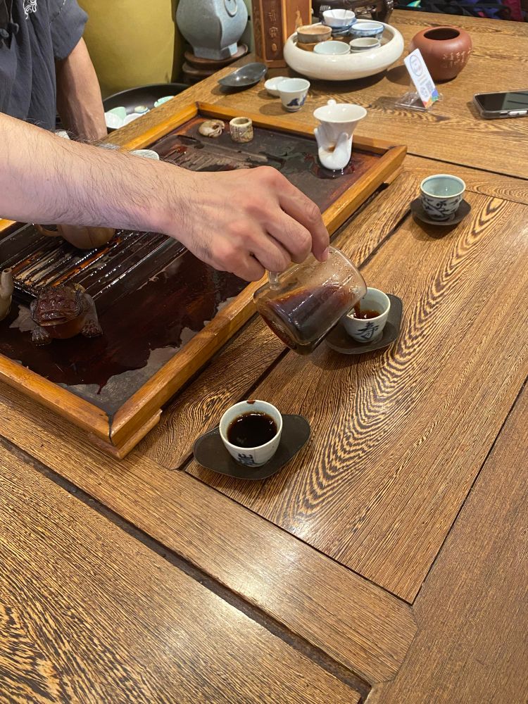 A persons hand pouring pu’er tea into the second in a row of three small tea cups. He reaches across the tea tray where a yixing tea pot can be seen. 