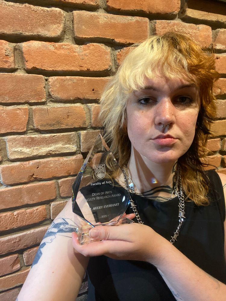 Pic of a white woman with calico hair looking into the camera. She holds a clear glass plaque in a tear drop shop with the university of British Columbia logo that reads faculty of arts, dean of arts faculty research award, Dr Avery everhart. Her shirt is black and sleeveless and she wears two silver necklaces. She stands in front of a brick wall. 