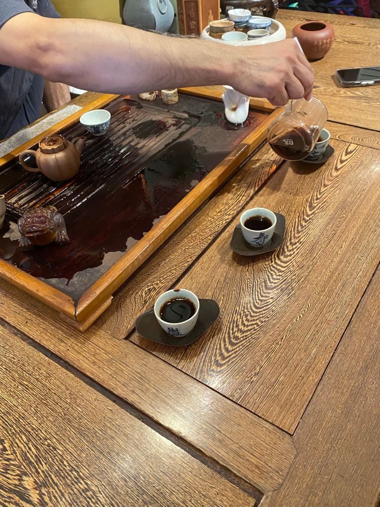 A persons hand pouring pu’er tea into the third in a row of three small tea cups. He reaches across the tea tray where a yixing tea pot can be seen. 