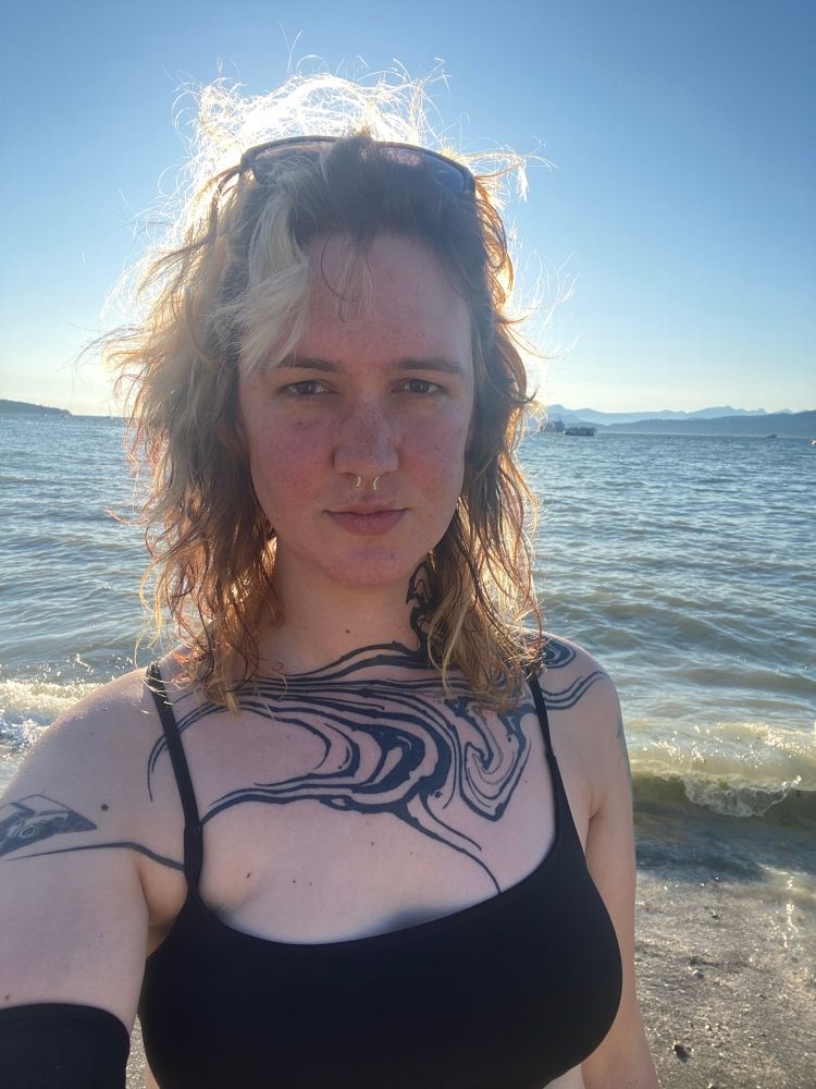 A white woman stands at the shoreline of a beach with mountains in the background.