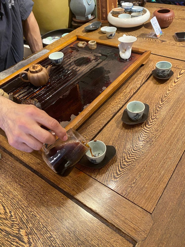 A persons hand pouring pu’er tea into the first in a row of three small tea cups. He reaches across the tea tray where a yixing tea pot can be seen. 