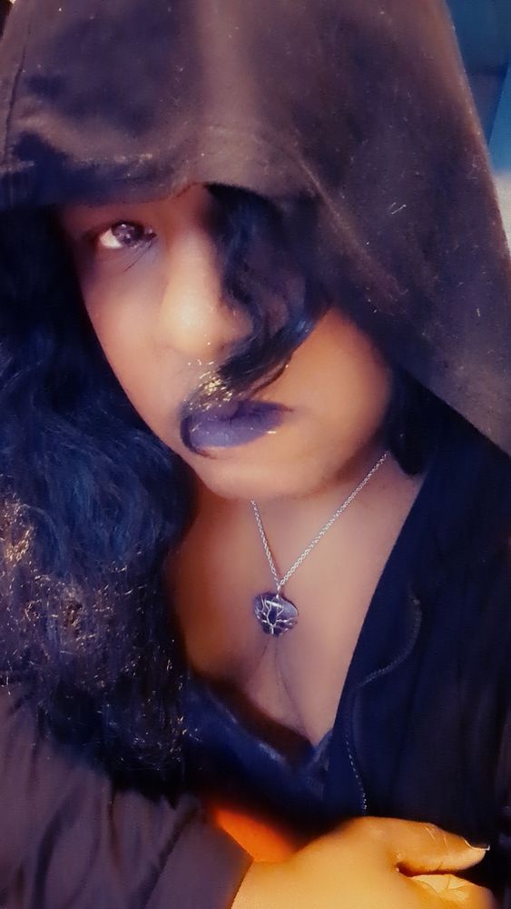 Portrait shot of Synn with long black hair, purple lipstick and a hood. Has major witchy vibes. 