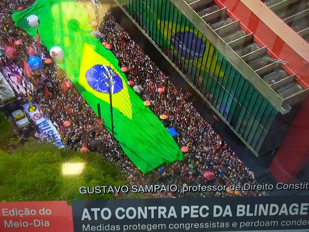 Foto aérea da av. Paulista. Uma multidão estica uma bandeira gigante do Brasil que é espelhada contra os vidros do prédio do MASP. É uma foto da TV cuja legenda diz "ato contra PEC da blindagem"