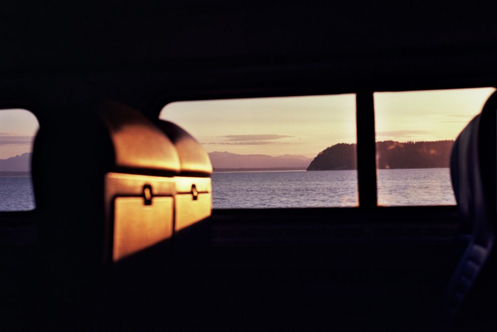 A color film photo, shot from the inside of a train at sunset. The interior of the car has been darkened out by contrast, leaving only the outlined shapes of the windows to show the Puget Sound at sunset. In the background, mountains fade into fog, while in the middle distance, a rocky outcropping rises from the water. The foreground coach seats are highlighted by bright orange evening sunlight. 