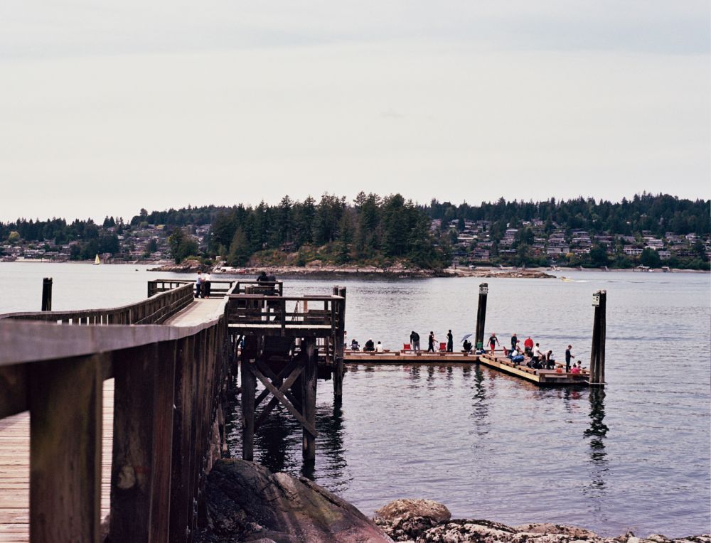 A color film photo of a dock floating on a lake. A small wooded island sits in the middle distance. In the background, a suburb rises up among the trees on the other shore. 