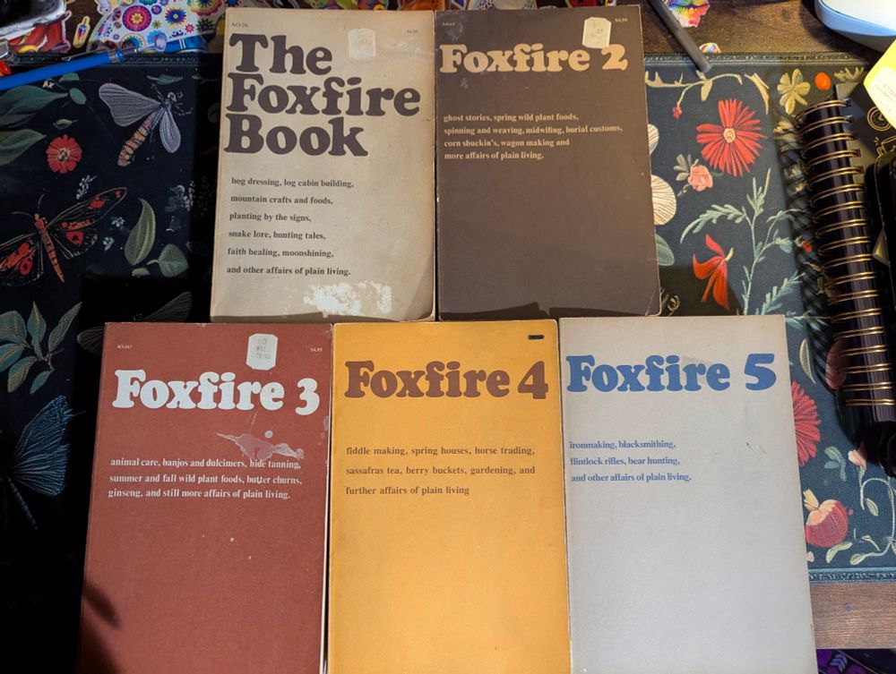 The Foxfire Book series: 1970s printings of books 1-5 out of 12 laid out on a desk with a dark floral desk mat
