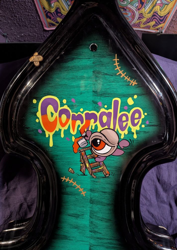A bright green to black burst style painted standup bass body with lettering in a creepy cartoon style of the Name Corralee being painted by an eyeball creature fashioned to look like Igor from the Frankenstein story. He is perched on a variegated leaf ladder with a broken leg that is about to fall over, and his paint can is spilling orange paint as it falls off the ladder. The illustration is surrounded by bronzed curved stitches on its top right and bottom left sides. 