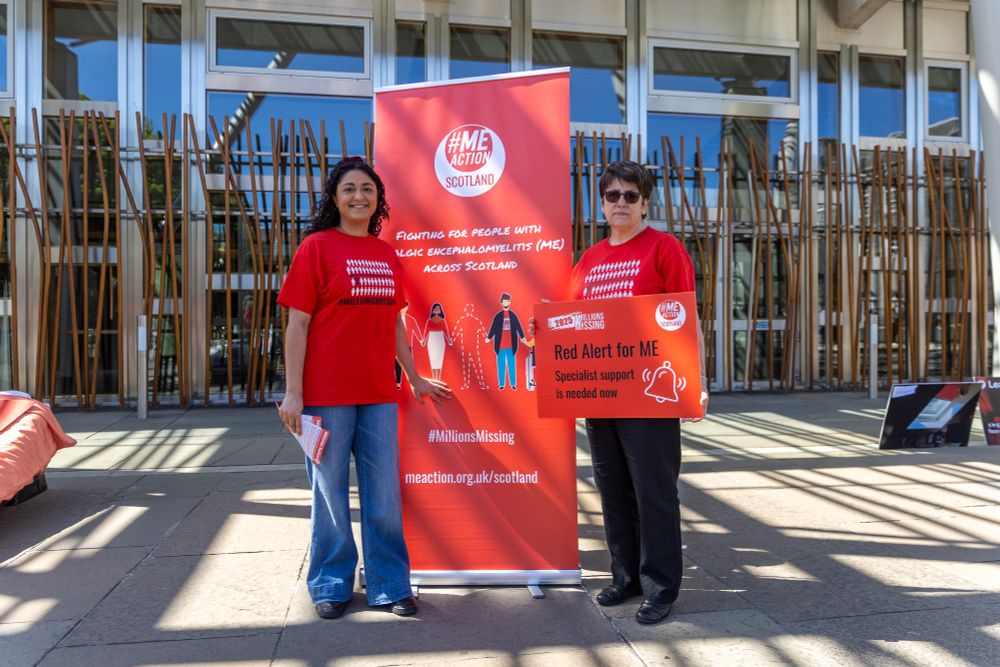 A photo of Tess White MSP standing with an ME Action Scotland volunteer outside the Scottish Parliament. They are standing next to an ME Action Scotland banner and Tess White is holding a red board that says 'Red Alert for ME, specialist support is needed now' and wearing a red ME Action t shirt.