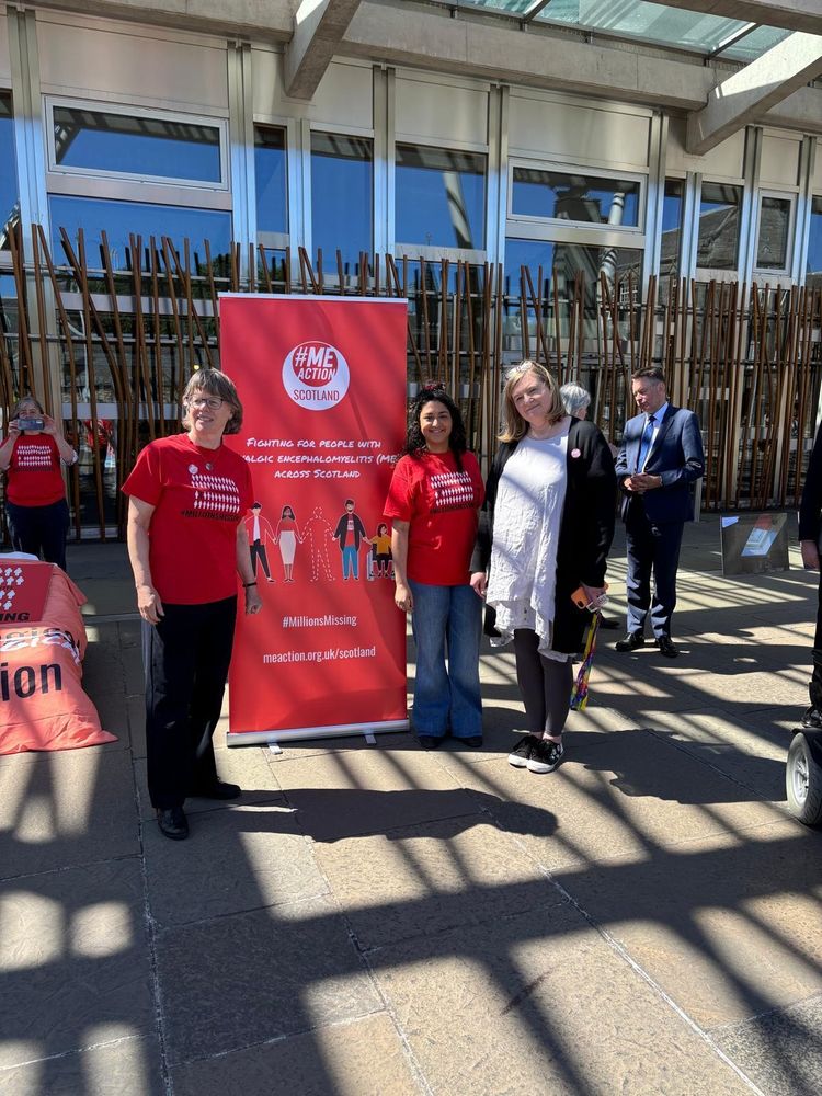 A photo of two ME Action Scotland volunteers standing with the Minister for Public Health and Women's Health, Jenni Minto, around an ME Action Scotland banner outside the Scottish Parliament.