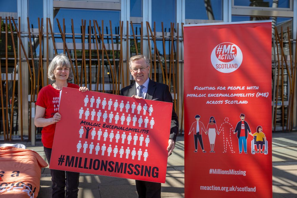 A photo of Alexander Stewart MSP standing with an ME Action Scotland volunteer outside the Scottish Parliament. They are holding a Millions Missing sign and standing next to an ME Action Scotland banner.