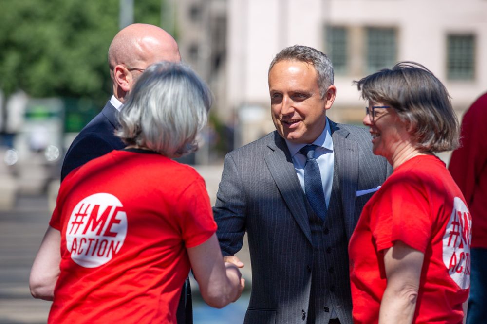A photo of Alex Cole-Hamilton MSP shaking hands with an ME Action Scotland volunteer outside the Scottish Parliament at the Millions Missing Scotland demonstration. Another ME Action Scotland volunteer and another man who is mostly obscured are standing with them.