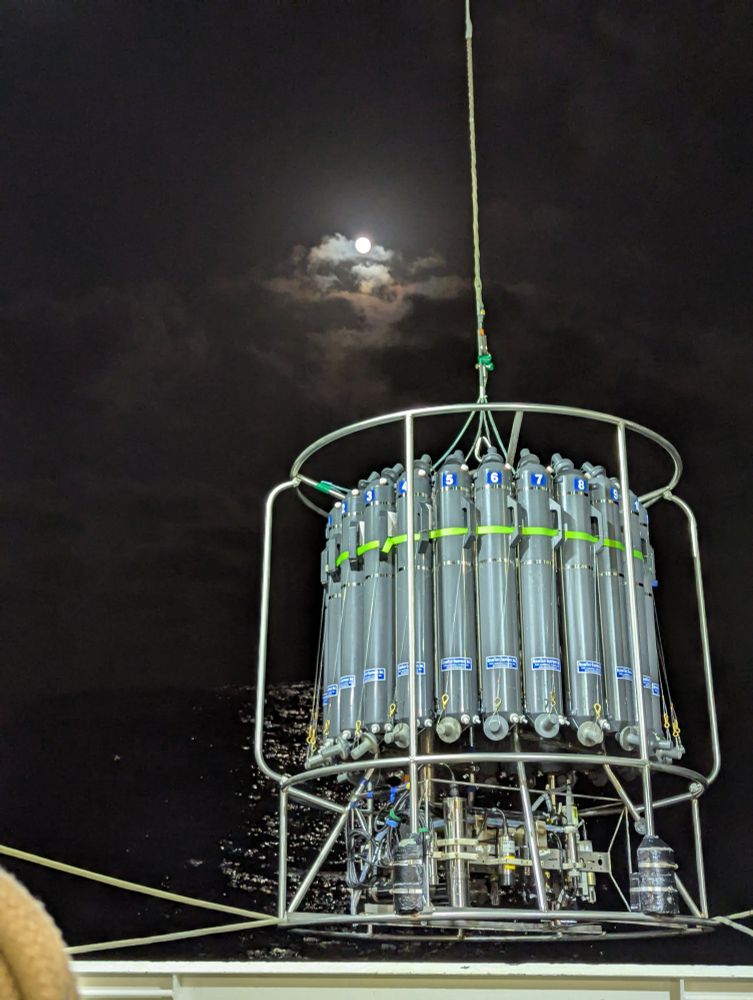 The CTD rosette hovers over the side of the R/V Atlantic Explorer in preparation for deployment at night for a diel sampling time point. Moonlight filters through clouds and reflects off the surface of the dark ocean. Photo by Claire Garfield.