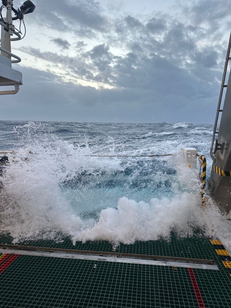 A wave crashes on the deck of the RV Atlantic Explorer during a storm, a rough sea streaked with white water and a stormy, grey sky visible in the background. Photo by McKenzie Powers.