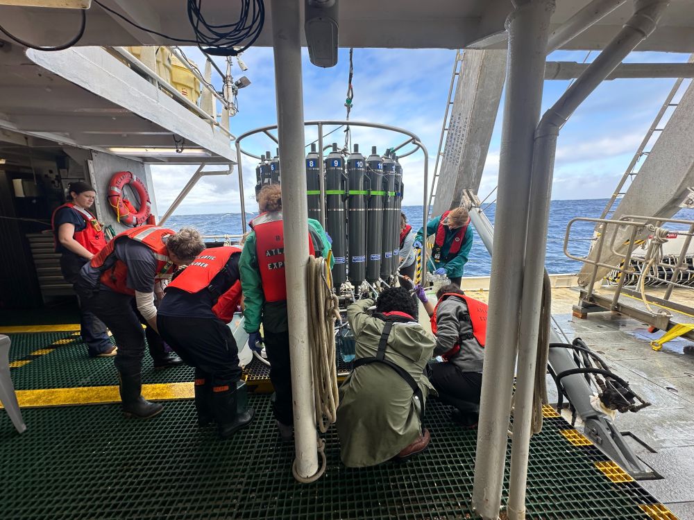 8 or so members of the research team gather around the CTD rosette on the deck of the RV Atlantic Explorer  to collect seawater for downstream sample processing. Photo by Emily Hu.