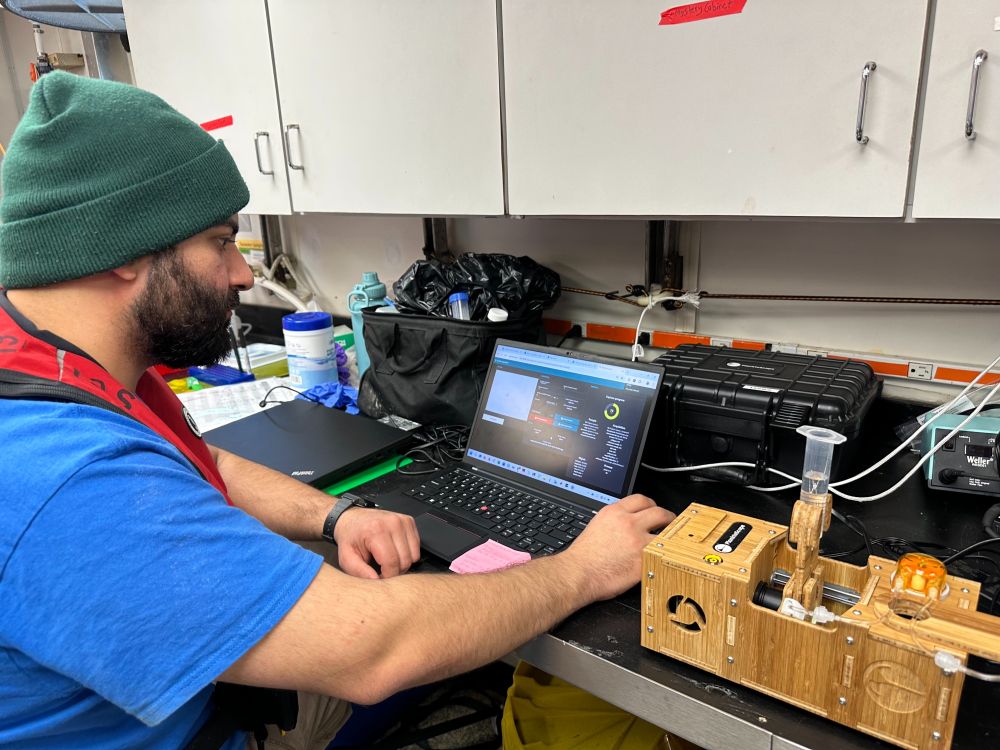 Dr. Loay Jabre runs the Planktoscope on the R/V Atlantic Explorer during the 2025 C-CoMP March Cruise. Loay collects images and videos of plankton on a laptop computer that is situated to the left of the Planktoscope on a lab bench. A sample is loaded onto the Planktoscope. Photo by McKenzie Powers. 