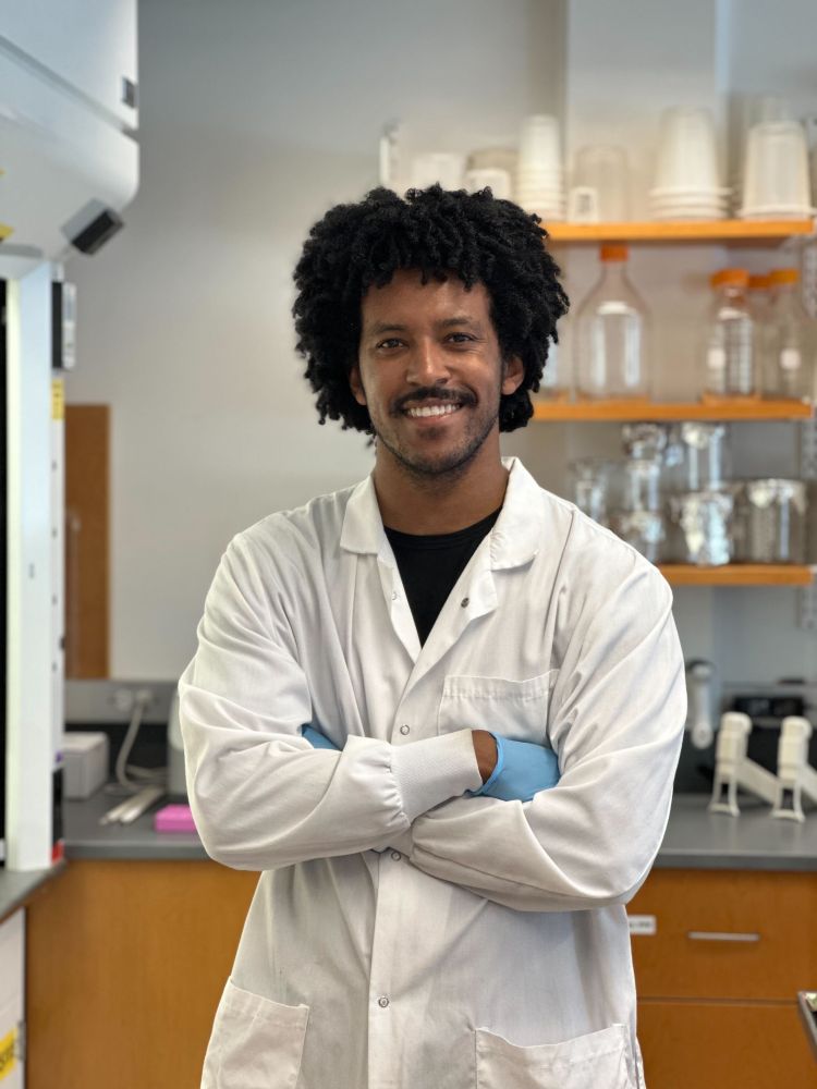 Headshot of Ben Acosta wearing a lab coat inside a laboratory with a lab bench and shelves of glassware in the background. Photo by Anna Chen. 