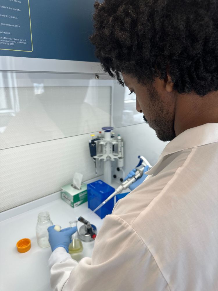 Ben prepares to pipette into a flask containing a light green/yellow solution within a culture hood. Photo by Anna Chen.