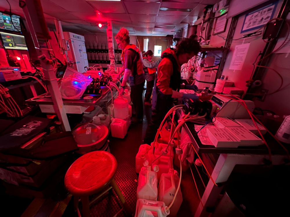 Claire Garfield, Sonya Dyhrman, and Arianna Krinos filter seawater collected during a diel time point in one of the labs on the R/V Atlantic Explorer. The room is bathed in red light (instead of bright white overhead lights) to minimize light exposure to seawater microbes that were collected at night. Photo by McKenzie Powers.