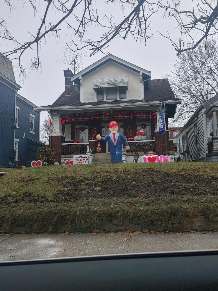 In front of a suburban house stands an ugly blow-up doll Trump among Valentines decorations.