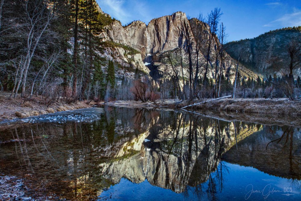 The early morning light hitting the mountain peaks of Yosemite National Park creating a perfect mirror reflection of the view in the perfectly still waters of the rive in the foreground.

The waterfall is practically frozen and a trickle of water cascades down the rocks creating subtle rainbow reflections in the showy air. 