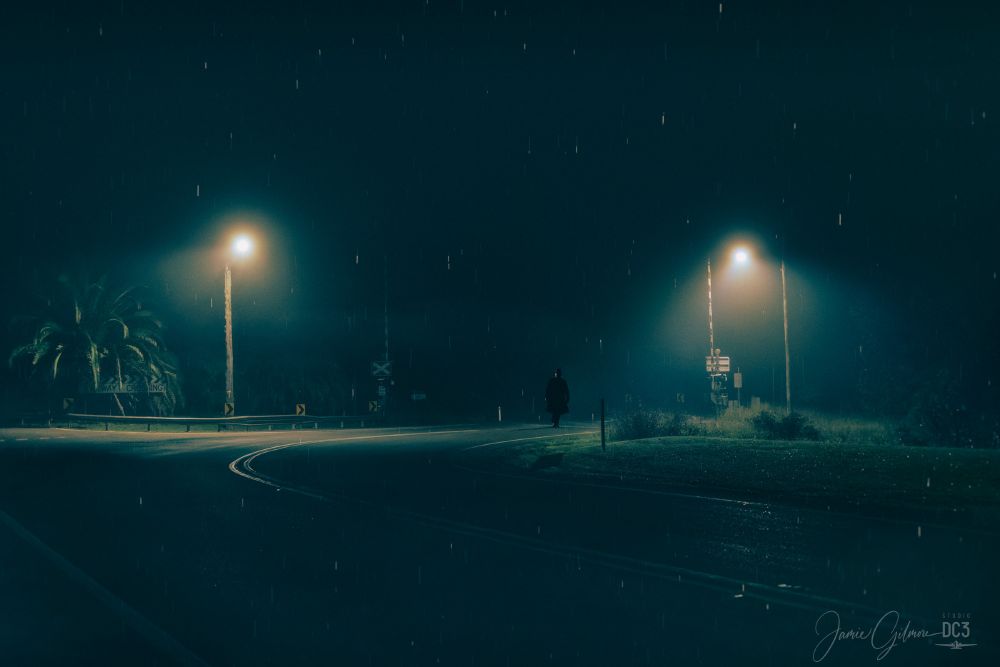 A man in a black suit with a long black coat and black fedora walks across the train crossing on a dark, rainy, foggy night. 

The road curves in a 'S' shape ahead, crossing the tracks briefly before twisting through the snaking curve towards town.

The light from the twin telegraph pole lights above the crossing are shrouded in fog, and cast an eerie glow just far enough to illuminate the Man in Black.

Photographed at the old rail crossing at the entryway to the City of Cessnock on the edge of Hunter Valley Wine Country, New South Wales, Australia.
