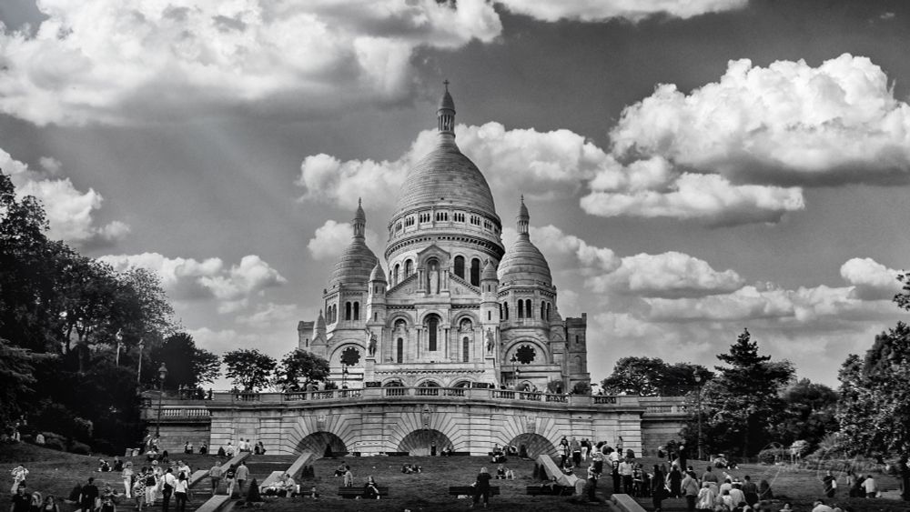 Timeless beauty in monochrome: The Sacré-Cœur Basilica - Paris.  
A moment frozen in time, where history and artistry meet the clouds.

This black and white photo of the famous Sacré-Cœur Basilica - Paris shows rays of light coming out from behind large fluffy white clouds towards the church. Many people walk the stairs or picnic on the grassy hillside leading to the church. 

While the church was designed in the Romana-Byzantine architectural style to make it look ancient, the building was built in 1914 and is only 110 years old. 

One of the key features of the design is the use of Chateau-Landon stone, which exudes calcite when it rains, causing the stone to become a bright white. 