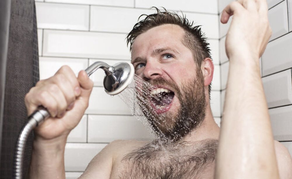 En skäggig man med hår på bröstet i duschen, där han ser ut att sjunga och använda duschmunstycket som mikrofon.