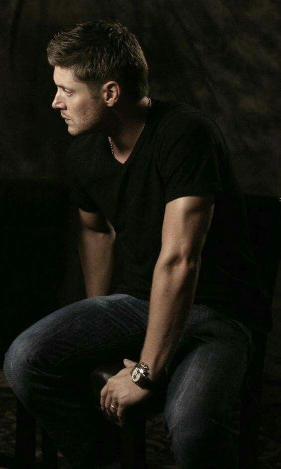 Jensen Ackles in a black t-shirt sits on a stool, facing away, with a focused expression, showcasing muscular arms and a casual style.