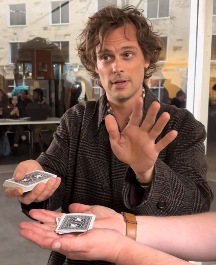 Matthew gray gubler in a checkered coat holds a deck of cards while gesturing towards another person's outstretched hand.