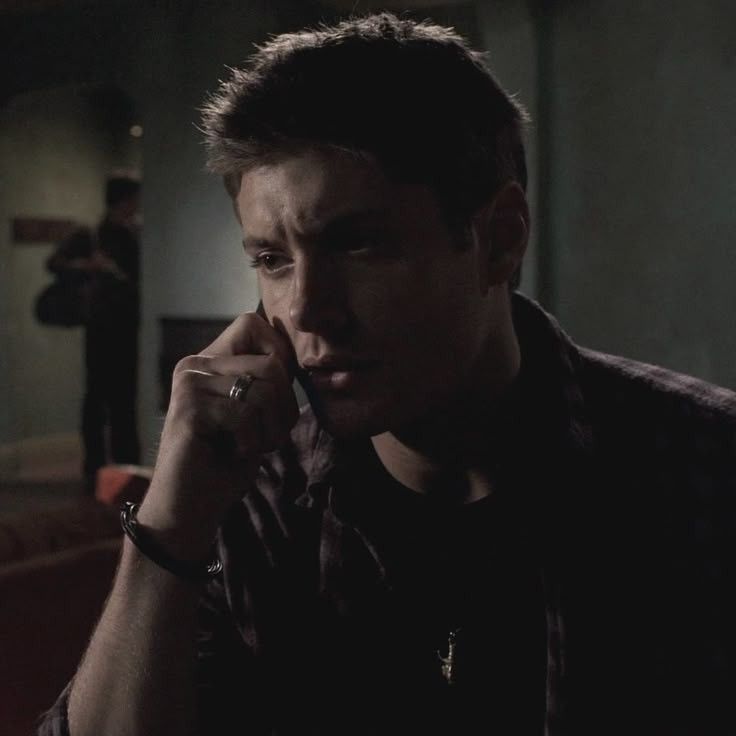 A dimly lit scene with Dean Winchester (Jensen Ackles) sitting in a contemplative pose, wearing a plaid shirt and holding a phone in Supernatural 