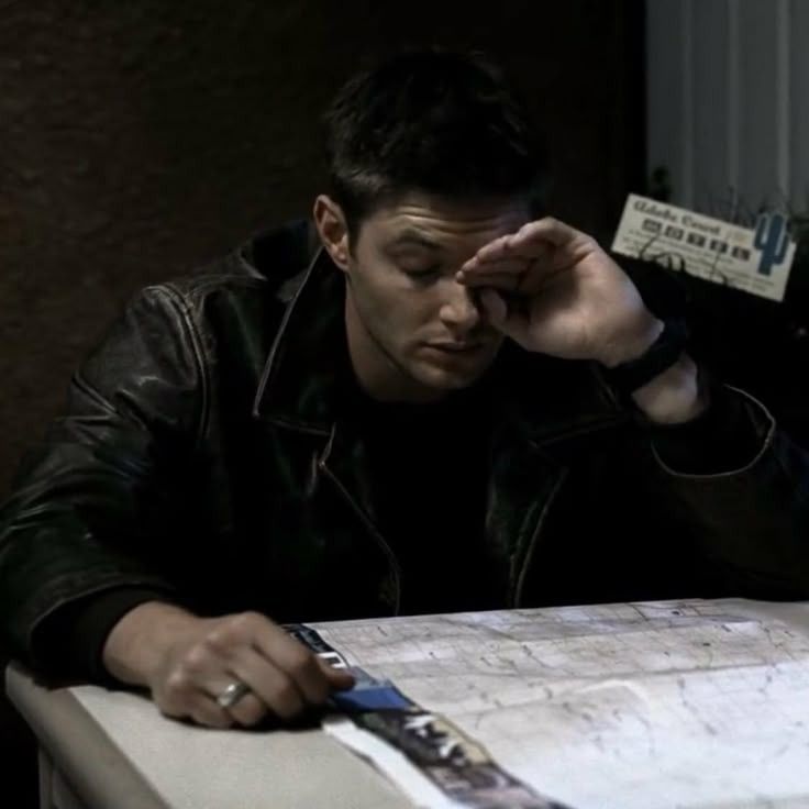 Dean Winchester (Jensen Ackles) in a leather jacket sits at a table, studying a map, rubbing his eyes in tiredness in supernatural 